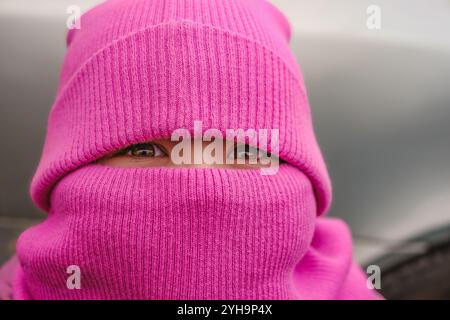 Un bambino con un berretto a maglia rosa, che copre la maggior parte del viso, sorride con occhi luminosi, pronto per un'accogliente giornata invernale. Foto Stock