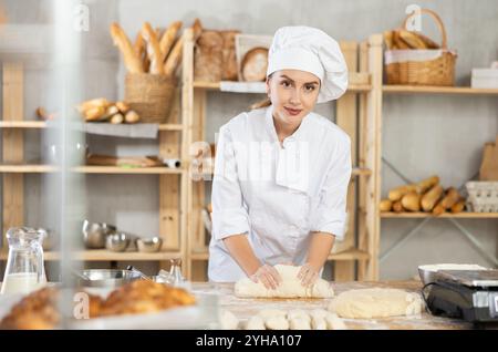 La panettiera femminile si trova al banco di lavoro, impastando e modellando l'impasto per fare pane, croissant e baguette Foto Stock