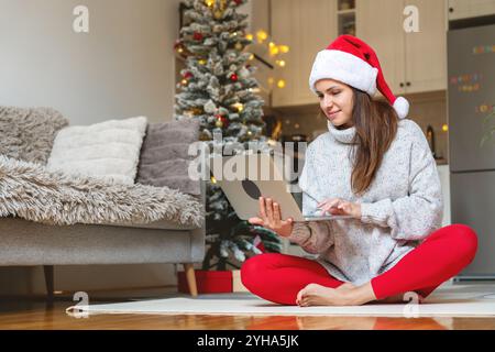 Donna con cappello di Babbo Natale siede sul pavimento in posa di mezzo loto e utilizza il suo laptop. Saldi della stagione invernale e shopping online. Foto Stock