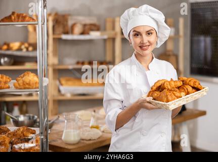 Ritratto di una giovane ragazza felice, fornaio in uniforme bianca e cappello da fornaio che lavora in panetteria, tenendo un vassoio con prodotti da forno freschi in cucina Foto Stock