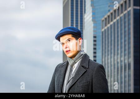 Un uomo vestito con un cappotto grigio e un berretto piatto blu guarda attentamente, con alti edifici moderni che torreggiano dietro di lui. Il cielo coperto aggiunge un atmos lunatico Foto Stock
