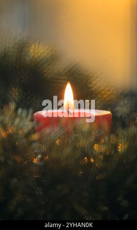 Candela rossa che brucia pacificamente nel bel mezzo di una corona di Natale, con luce del tramonto che arriva attraverso una finestra dietro di essa Foto Stock
