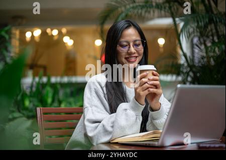 Una bella e rilassata donna asiatica in un intimo maglione si gode un caffè mentre lavora a distanza da una caffetteria. bevete, gente, stili di vita Foto Stock