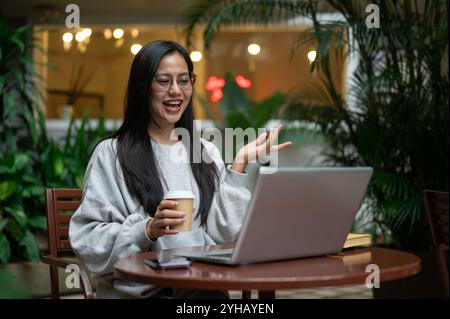 Una donna asiatica attraente e positiva con un maglione accogliente e occhiali da vista ha un incontro online con il suo team, studia online o tutor a distanza sulla sua famiglia Foto Stock