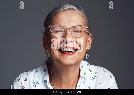 Donna anziana, occhiali e felice in studio per un atteggiamento positivo, fiducia e gioia su sfondo grigio. Persona anziana, sorriso e allegra per Foto Stock