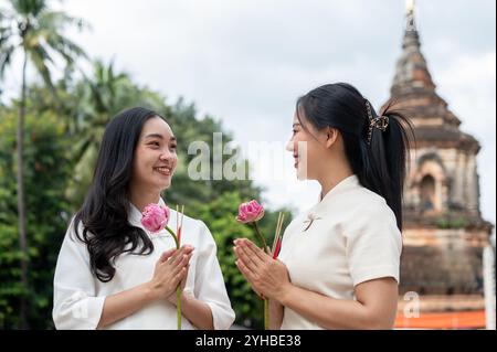 Due belle donne asiatiche con abiti tradizionali thailandesi del nord-Lanna si stagliano in un tempio, tenendo in mano lotuses rosa e sorridendo l'una con l'altra. Cultura thailandese, Foto Stock