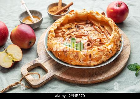 Una crostata di mele appena sfornata siede su un rustico tavolo di legno, guarnita con mandorle e menta, accompagnata da mele rosse e una piccola ciotola di miele, inviti Foto Stock