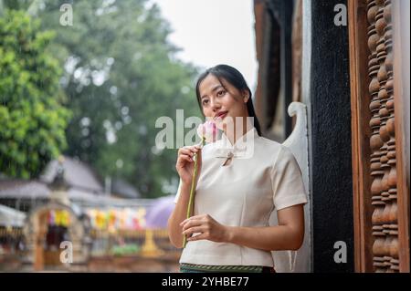 Una bella donna asiatica in un tradizionale abito Thai-Lanna si erge in un tempio, tenendo un loto e sorridendo lontano dalla macchina fotografica. Cultura tailandese, buddista Foto Stock