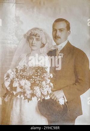 Jugoslavia - 1919: Ritratto di nozze di una bella coppia di sposi novelli in piacevoli tonalità seppia; sposa in abito da sposa bianco, con velo bianco che regge un grande bouquet di fiori bianchi, sposo con un baffo in piedi dietro di lei indossa un abito scuro con un fiore nel suo risvolto, cravatta con fiocco bianco e tenendo in mano un cappello da bowler e guanti da whote Foto Stock