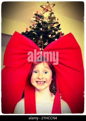 Young redheaded girl looking silly with a giant red bow on her head in front of a Christmas tree - Fair Oaks California Stock Photo