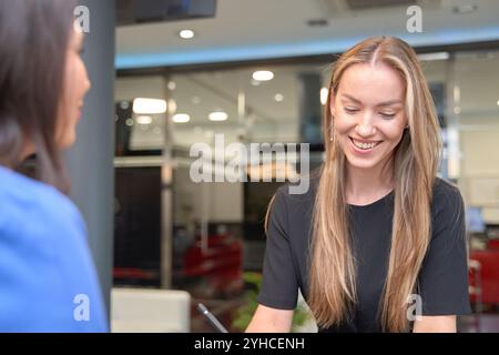 Una donna in abito blu sta scrivendo su un tablet alla reception di una clinica o di un dentista. Una donna in camicia nera gli sorride. Foto Stock