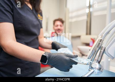 giovane dentista latino vestito di blu, dettaglio che toglie utensili da sacchetti sterili, sfondo dello stand del dentista con il cliente seduto, modalità irriconoscibile Foto Stock