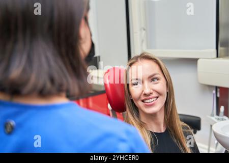Una donna con una camicia nera siede sulla sedia di un dentista mentre una donna con una camicia blu le parla Foto Stock