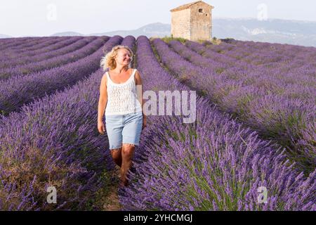 Giovane donna bionda sorridente che si gode una passeggiata nei filari di fiori di lavanda con l'antica fattoria tradizionale in pietra durante il tramonto a Valensole Fields i. Foto Stock