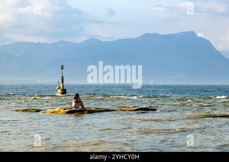 Sirmione, Italia - 21 settembre 2024: Una donna si rilassa sulle rocce del lago, godendosi la tranquilla vista sul Lago di Garda Foto Stock