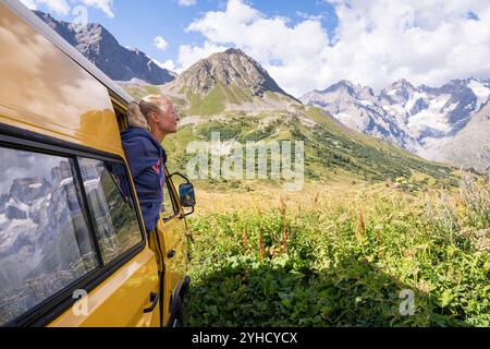 Giovane donna bionda che gode della vista sulle montagne nel suo camper giallo retro nel parco nazionale di Ecrins, in Francia Foto Stock