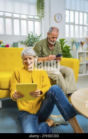 Una coppia di anziani che utilizza un tablet digitale e un telefono cellulare trascorre del tempo rilassato a casa Foto Stock