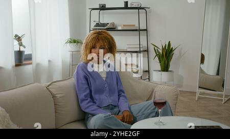 Calma rilassata triste ragazza afroamericana da sola donna tranquilla donna tranquilla cattiva abitudine femmina nel soggiorno di casa appartamento che tiene un bicchiere di vino rosso gustando Foto Stock