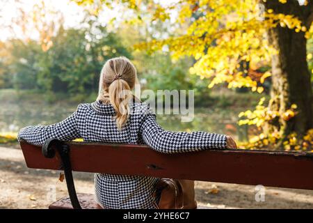 Donna che si rilassa sulla panchina del parco in autunno e si gode una giornata di sole. Donna bionda che indossa un cappotto pied de poule Foto Stock