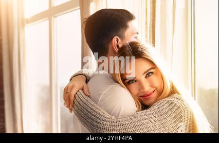 Giovane ragazzo e donna che si abbracciano vicino alla finestra in una stanza illuminata dal sole Foto Stock