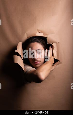 La testa di un uomo si stacca e guarda attraverso un foro di carta con le mani che spingono la carta da parte. Ritratto in studio. Foto Stock