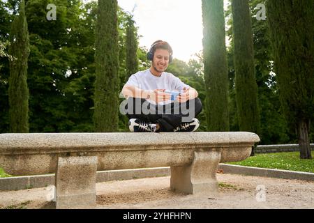 Un uomo è seduto su una panchina con la testa abbassata e un cellulare in mano. Indossa le cuffie e si concentra sul telefono. Foto Stock