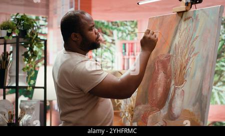Artista afroamericano che usa la pittura ad acquerello e mescola i colori su tavolozza per la sua creazione artistica in un laboratorio di belle arti. Uomo che dipinge elementi rustici su tela, processo creativo. Telecamera A.. Foto Stock