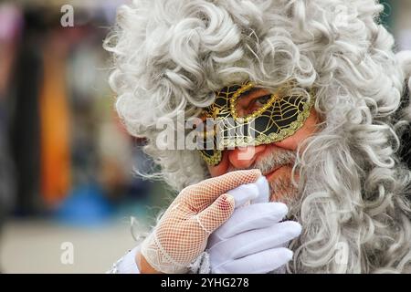 Primo piano di un uomo anziano vestito con un elaborato costume di carnevale, indossando una parrucca bianca riccia e una maschera nera con motivi dorati a rete. Foto Stock