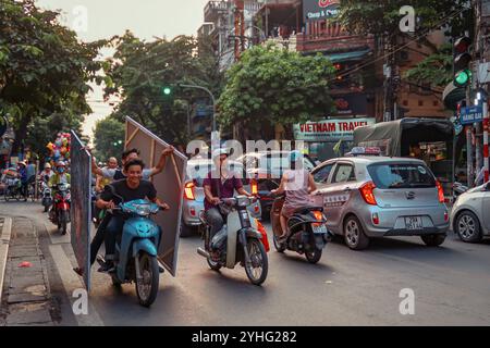 I motociclisti che trasportano grandi e ingombranti oggetti navigano attraverso le strade trafficate di Hanoi, Vietnam, evidenziando il trambusto quotidiano della città Foto Stock