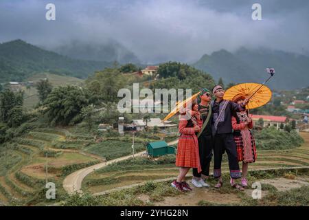 Un gruppo di amici vestiti con abiti tradizionali vietnamiti, in posa per un selfie con ombrelli gialli su una collina, che si affaccia sui campi terrazzati. Foto Stock