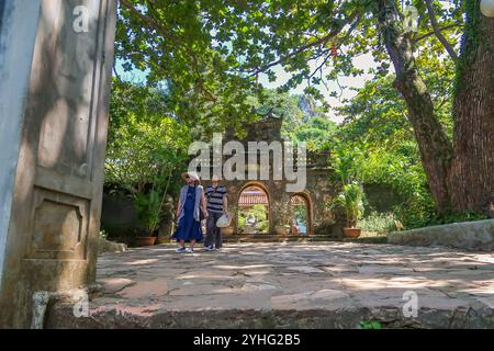 I visitatori camminano attraverso l'ingresso di uno storico tempio vietnamita circondato da vegetazione lussureggiante e grandi alberi, catturando l'atmosfera tranquilla. Foto Stock