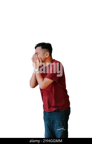 Un uomo con una camicia rossa e un jeans copre il viso con entrambe le mani, mostrando angoscia o frustrazione. Isolato su uno sfondo bianco, la sua postura suggerisce Foto Stock