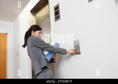 Donna in tuta che preme il pulsante dell'ascensore con molti documenti Foto Stock