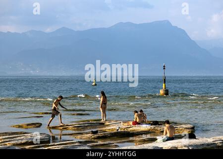 Sirmione, Italia - 21 settembre 2024: Le persone si rilassano sulle rocce del lago, godendosi la tranquilla vista sul Lago di Garda Foto Stock