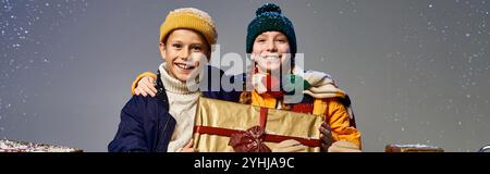 Due bambini allegri vestiti con comodi abiti invernali festeggiano con un regalo splendidamente avvolto. Foto Stock