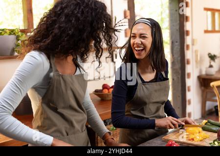 Due amiche multirazziali che ridono e preparano verdure insieme in un'accogliente cucina, a casa Foto Stock