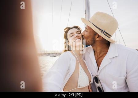 Una coppia sorridente scatta un selfie su una barca a vela durante una giornata di sole. L'uomo bacia dolcemente la guancia della donna, catturando un'atmosfera gioiosa e amorevole su t Foto Stock