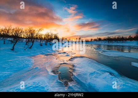 La vibrante alba proietta un caldo bagliore su un paesaggio innevato sul fiume, dove alberi sagomati incorniciano la scena. L'acqua riflette le tonalità arancio e blu Foto Stock