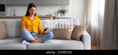 Giovane donna che si diverte con il suo smartphone mentre si rilassa su un comodo divano di casa Foto Stock