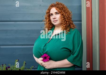 Plus size Woman con capelli ricci rossi in abbigliamento verde mantiene il fiore sullo sfondo blu della vivace parete, bellezza e sicurezza all'aperto Foto Stock