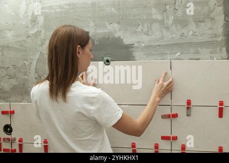 Donna che affianca le pareti del bagno, applica l'adesivo su una piastrella e posiziona la piastrella in ceramica nella giusta posizione. Processo di installazione delle piastrelle nel bagno Foto Stock