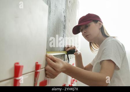 Donna che piastrella le pareti del bagno, utilizzando un metro a nastro per misurare la lunghezza. Processo di installazione delle piastrelle nel bagno passo dopo passo. Miglioramento fai da te Foto Stock