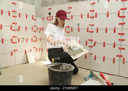 Donna che affianca le pareti del bagno, applica l'adesivo su una piastrella e posiziona la piastrella in ceramica nella giusta posizione. Processo di installazione delle piastrelle nel bagno Foto Stock