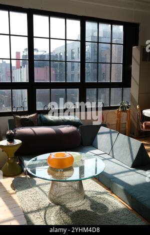 Soggiorno moderno in stile loft con grandi finestre industriali, tavolino da caffè in vetro, arredi eleganti e comode poltrone con luce naturale Foto Stock