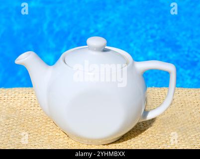 Bollitore con tè sullo sfondo della piscina. Tè alle erbe rinfrescante. Pentola in ceramica bianca. Foto Stock