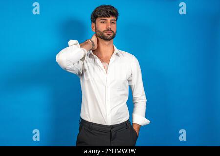 Uomo d'affari sicuro che posa con la mano sul collo, indossa camicia bianca e pantaloni scuri sullo sfondo blu brillante Foto Stock