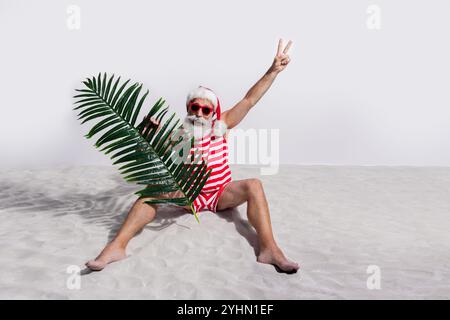 Foto di babbo natale con la barba indossa un costume da bagno a righe goditi le vacanze estive isolate sullo sfondo di una spiaggia di sabbia bianca Foto Stock