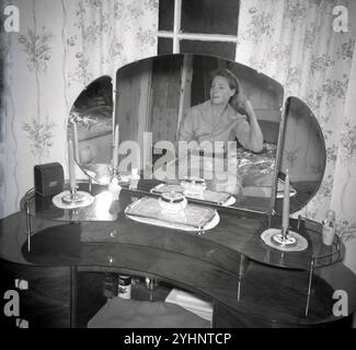 anni '1960, storica, all'interno di una camera da letto, una donna seduta su un letto, che si lavava i capelli, il suo riflesso visto nello specchio di un elegante toletta. Foto Stock