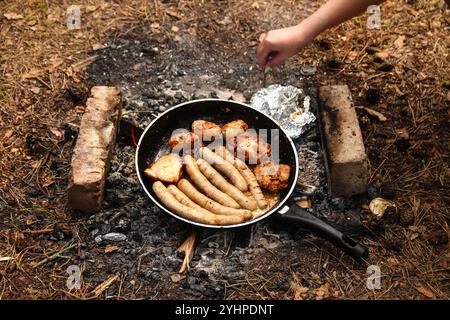 Una mano che regge una padella con salsicce che sfrigolano su un falò nella foresta. Le salsicce stanno cucinando sopra le fiamme, aggiungendo al rustico Foto Stock