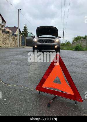 Un veicolo con cofano aperto è bloccato su una strada rurale. Viene impostato un triangolo segnaletico riflettente, che indica una situazione di avaria della vettura, circondato da r Foto Stock